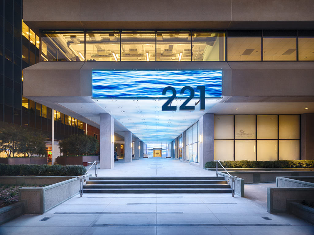 Exterior of modern office building, with a long digital canopy showing a water scene. The canopy lights up the street at night.