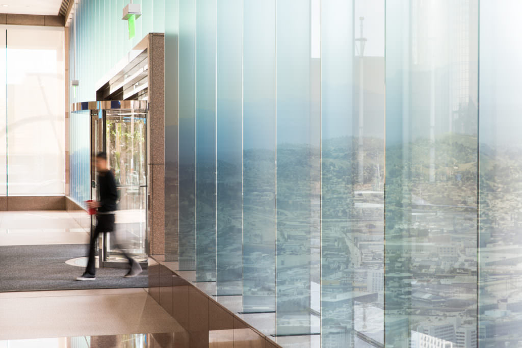 The modern office lobby at 1CAL Plaza in Los Angeles has vertical glass fins on the window on the right, which together make up a panoramic photo of the LA hills. A man speedily walks through the revolving door.